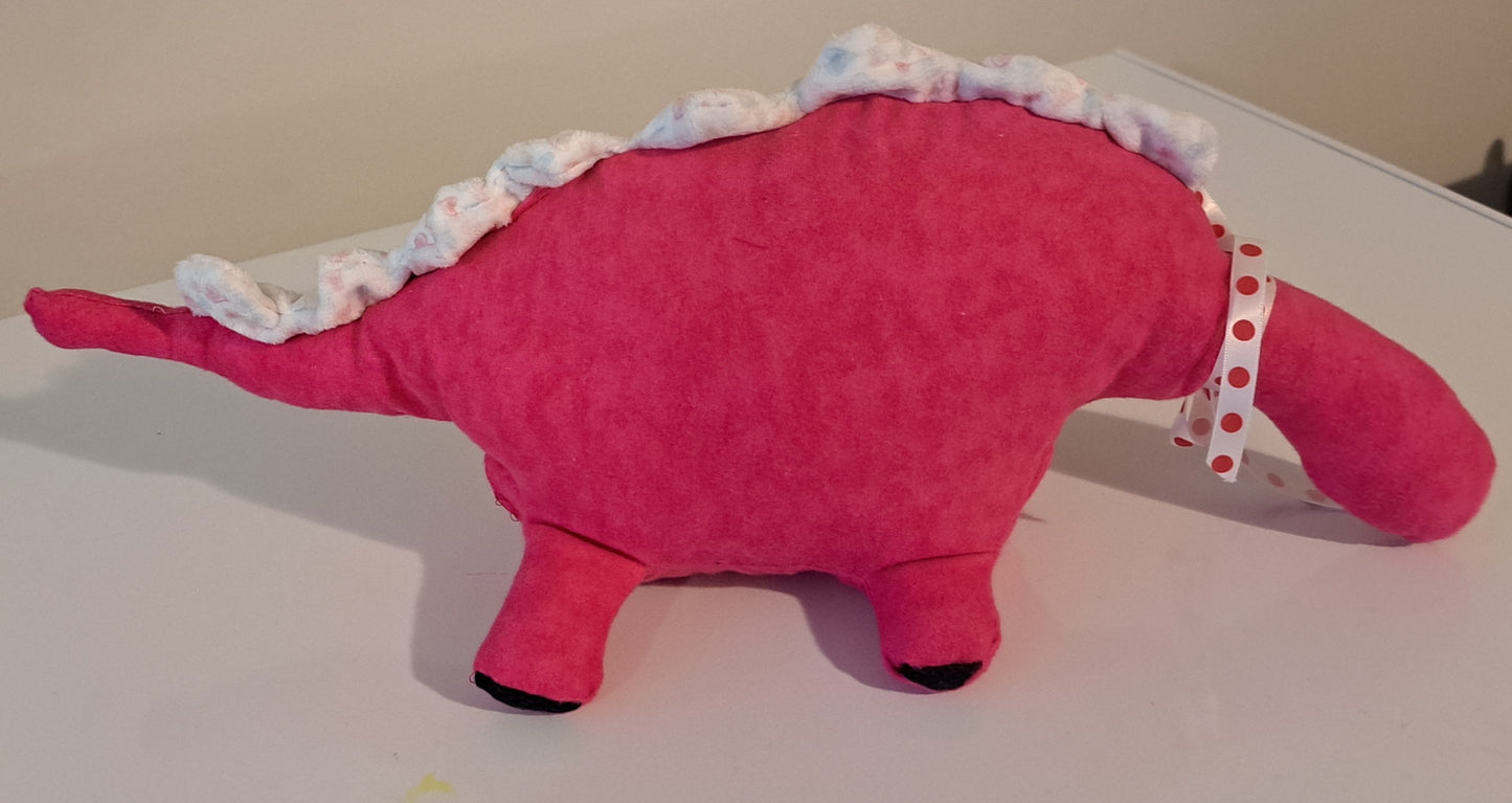 A pink plush dinosaur toy with a white and pink patterned ruffled tail and a red and white polka dot ribbon, resting on a light-colored surface.