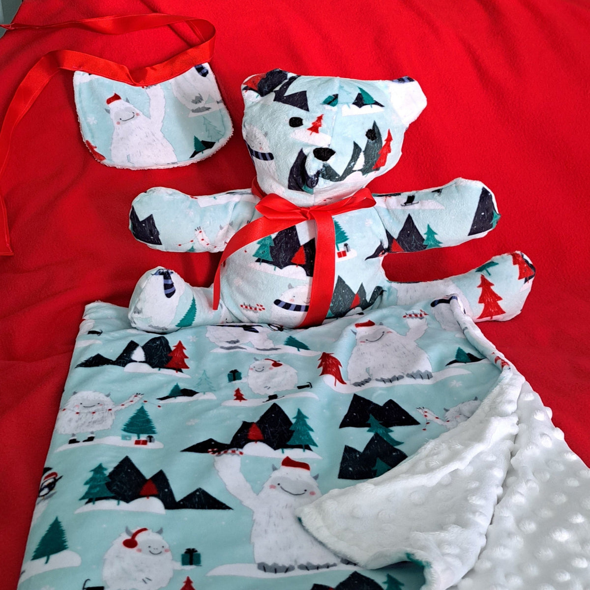 A handmade Christmas-themed baby gift set featuring a plush teddy bear, a matching pouch, and a soft blanket with a snowy landscape print, all decorated with red ribbons and laid out on a bright red fabric background.