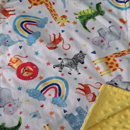 A close-up of a white plush blanket with a colorful safari animal print including a zebra, lion, giraffe, monkey, elephant, and rainbow, with a folded yellow minky fabric corner visible at the bottom right.