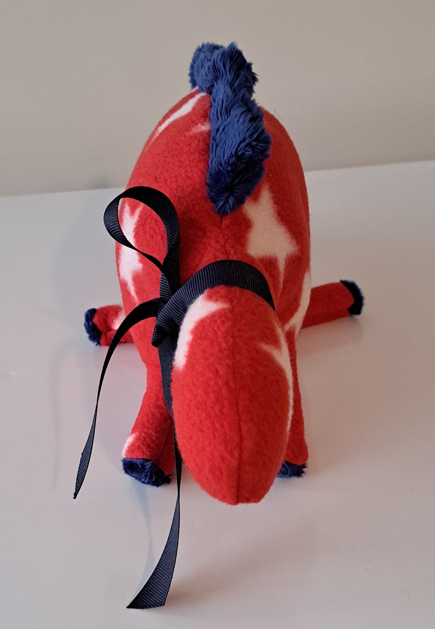 A red and white star-patterned plush toy with a blue mane and tail, featuring a long black ribbon hanging from its neck, set against a plain light background.