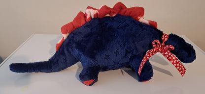 A plush dinosaur toy in dark blue with red and white accents, featuring a red and white frill along its back and a red patterned ribbon bow around its neck, resting on a white surface.