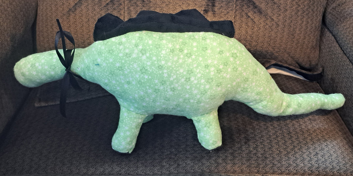 A light green dinosaur plush toy with a black mane and a black backpack, sitting on a brown textured couch.