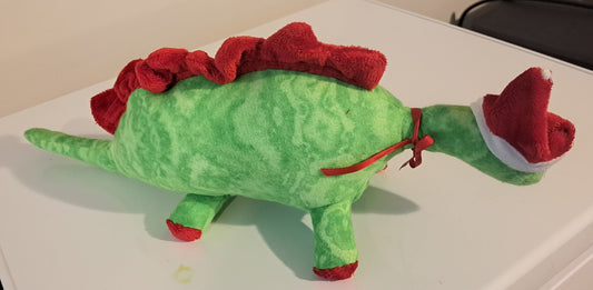 A green plush dinosaur toy with a red ruffled back and a red Santa hat on its head, sitting on a white surface.