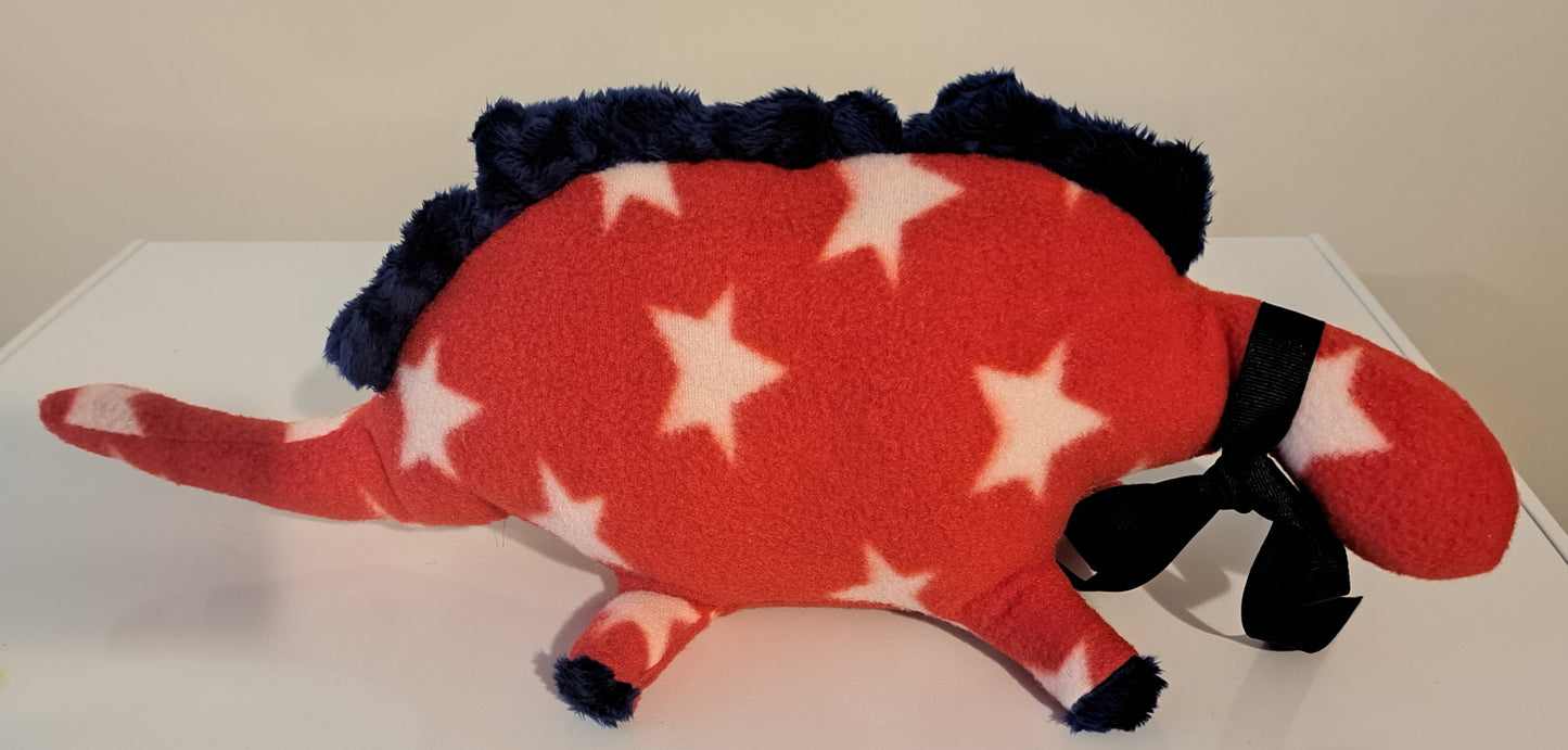 A plush dinosaur toy in a red fleece body with white stars, dark navy blue spiked back, and black ribbon accents, resting on a white surface against a beige wall.