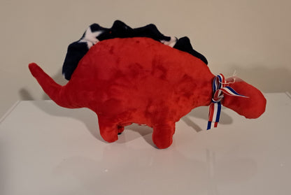 A red plush dinosaur toy with a black, white, and blue star-patterned tail and a red, white, and blue ribbon bow on its neck, displayed on a white surface against a beige wall.