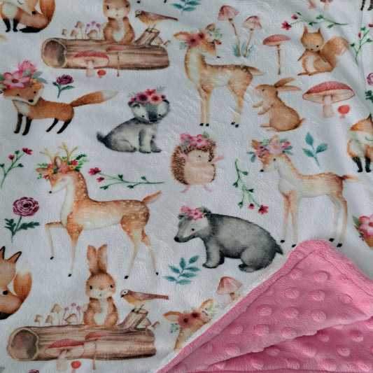 Handcrafted receiving blanket featuring a whimsical woodland animal print on a white background, with a folded pink minky dot fleece backing visible in the lower right corner.